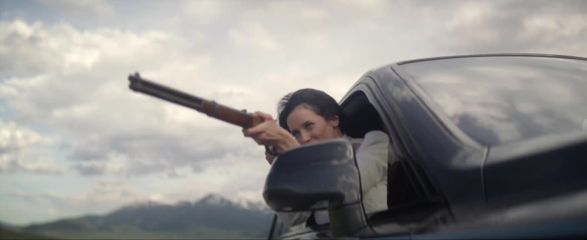 A woman leans out the passenger window of a moving vehicle and fires a rifle repeatedly