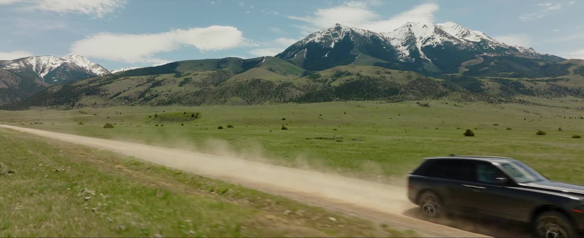 A black automobile is traveling on a rural dirt road with mountains in the distance