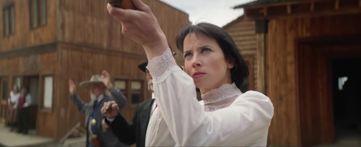 A woman carrying a rifle storms out of a building and walks to some men with their hands in the air and starts firing the gun at people