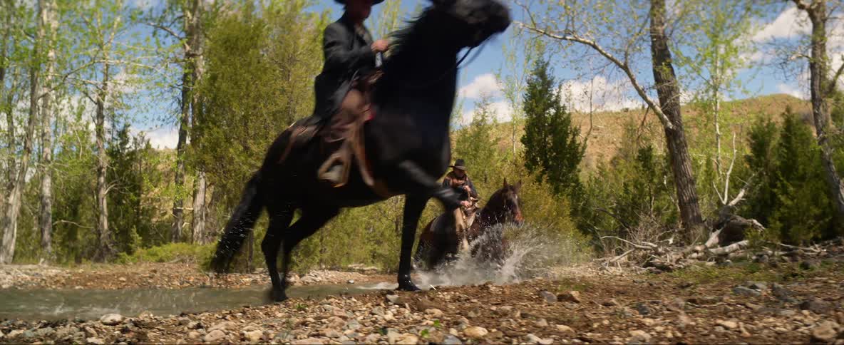A group of old time cowboys riding horses cross a river in the wilderness