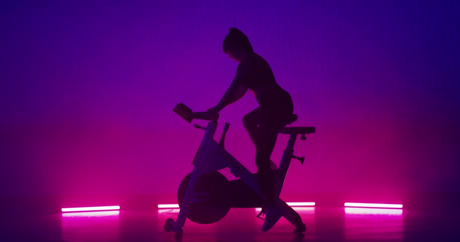 Woman with high ponytail approaches exercise bike and rides it against a pink and purple background