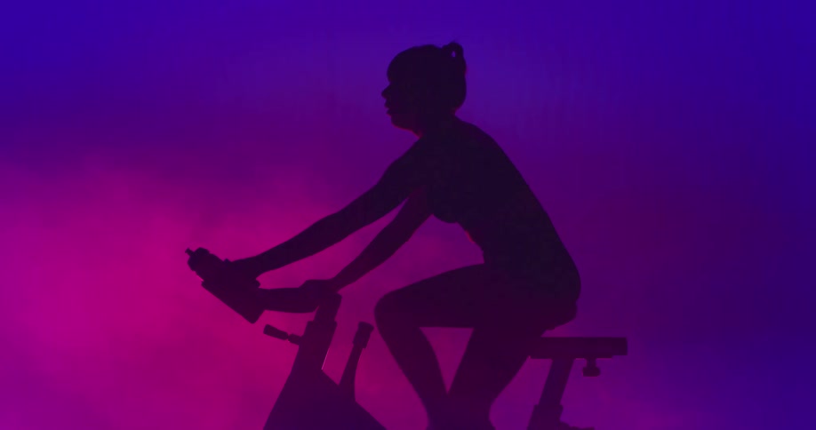 A person shown in silhouette on an exercise bike drinks from a sport bottle