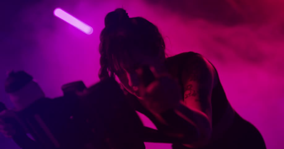 Young tattooed woman pedals intensely on a static bicycle una room full of smoke and colorful lights
