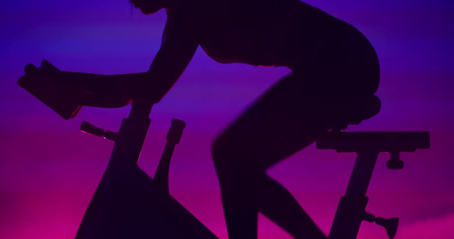 Silhouette of a woman pedaling vigorously as she rides an exercise bike with pink and purple smoke in the background