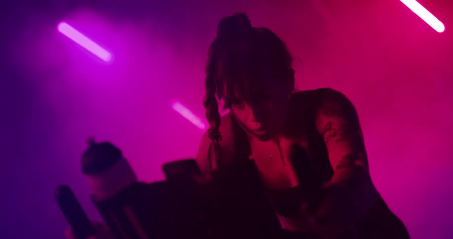 A woman is pedaling an exercise bike and there are lights and pink and purple smoke in the distance