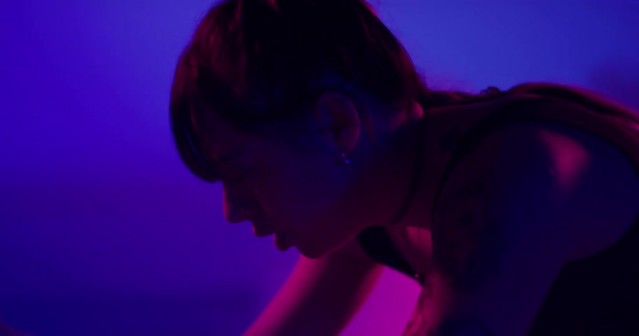 Woman exercises on stationary bike under colored lighting as white smoke rises in the background