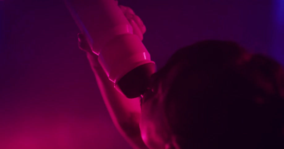 A woman is riding an exercise bike and takes a sip of water inside of a room with pink lights