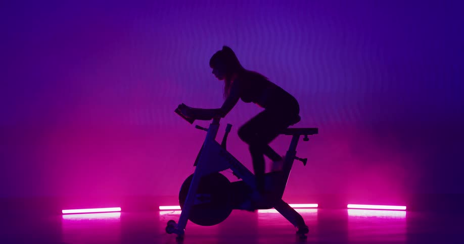 Silhouette of a woman riding an exercise bike on a stage with lights and pink and purple smoke in the background