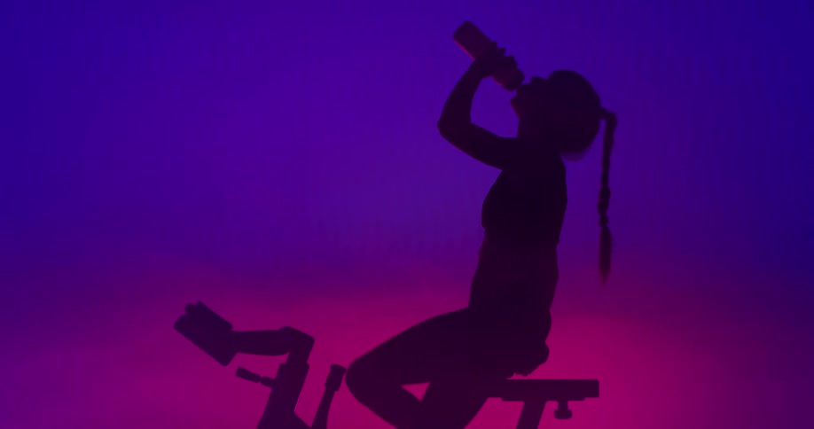 Woman with ponytail exercise bike is silhouetted against the led lights in the gym drinking water