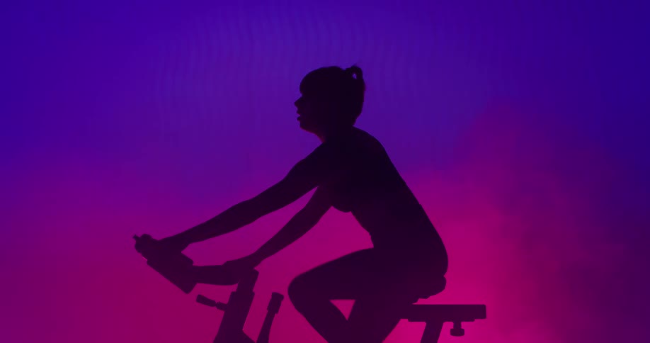 Woman with ponytail on exercise bike is silhouetted against the led lights in the gym