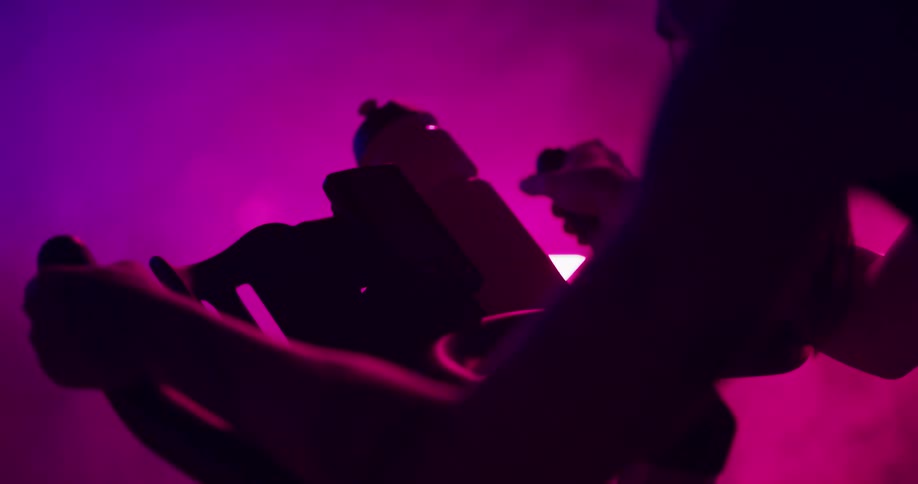 Woman exercises on stationary bike and takes drink from water bottle in room lit by colored lighting and filled with fog