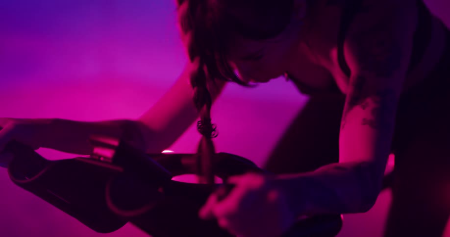 Woman pedals on stationary bike in room lit with colored lighting as smoke spreads in the background