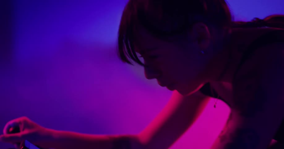 A woman is pedaling an exercise bike and there are lights and pink and purple smoke in the distance