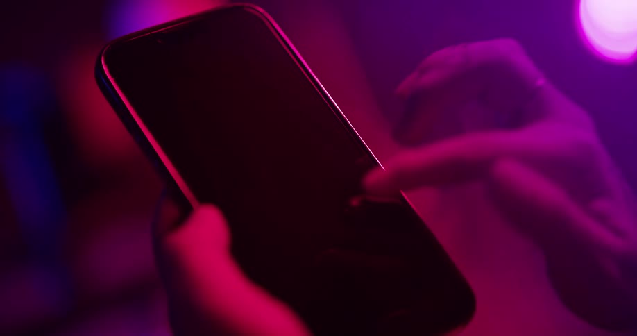 Close up of the tattoed hand of a woman gesturing on the screen of a smartphone under colorful lights