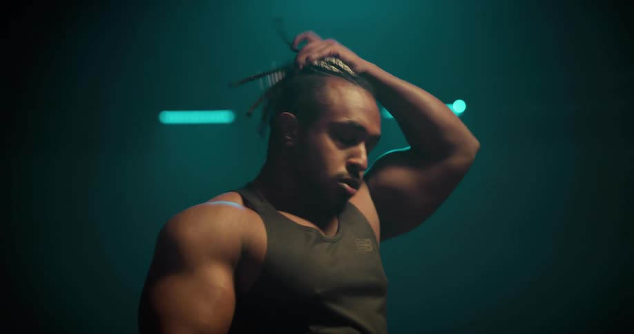 A muscular man paces and takes deep breaths while tying his hair in a dimly lit room