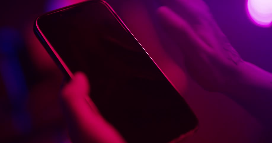 Close up of the tattoed hand of a woman gesturing over the screen of a smartphone under colorful lights