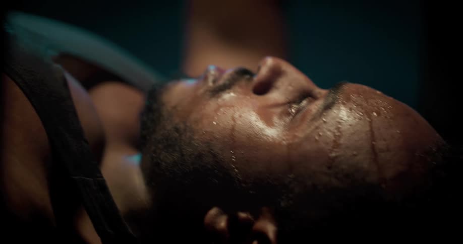 A sweating man lifts weights on a weight bench in a dimly lit room