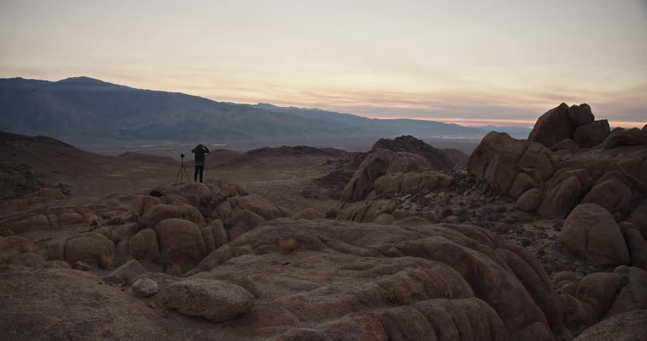 Photographer standing next to a tripod over a rocky hill in the desert at sunset