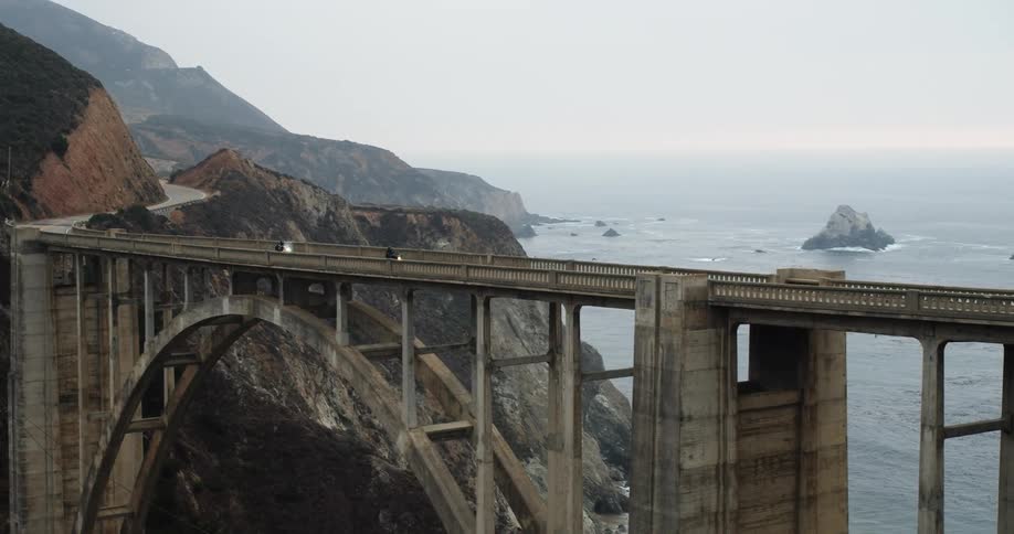Two people riding motorcycles cross a bridge in the mountains next to the ocean