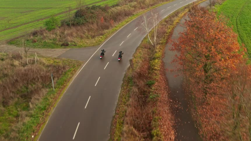 Two cyclists are riding motorcycles on a rural road lined with trees in beautiful fall colors