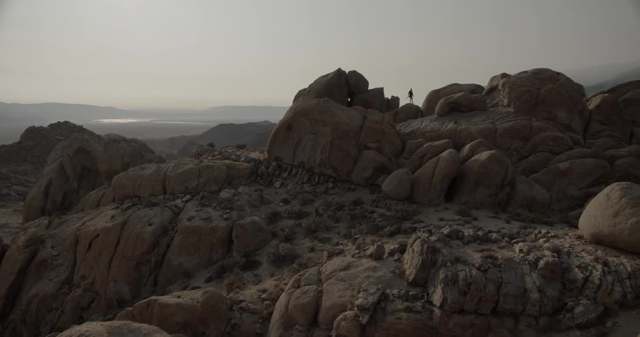 Silhouette of a person standing on top of a rock formation in the badlands with mountains in the distance under a hazy sky