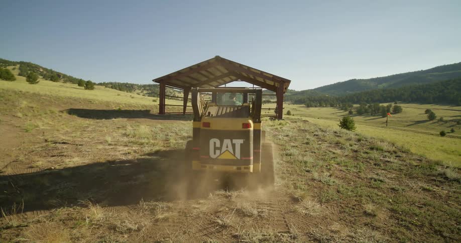 Male operator drives cat vehicle along ground toward structure in field
