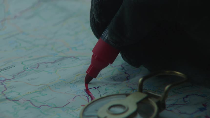 Close up of a gloved hand using a red felt pen to mark a route on a map next to a compass