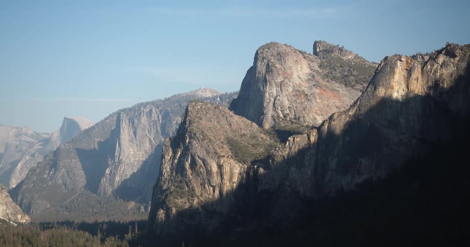 The sun shines brightly upon mountains with a forest in the valley under a blue sky