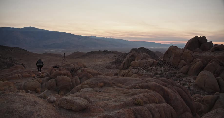A man is standing on rocks in the desert badlands looking at the mountains and he leaps from rock to rock to his camera on a tripod set up on a group of boulders