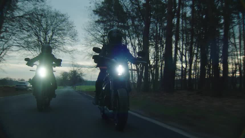 Two cyclists are riding motorcycles on a rural road lined with trees