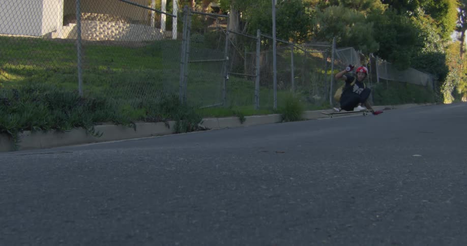 Skateboarder crouches and touches asphalt while riding down sloping street past fenced yards