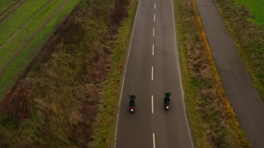 A pair of motorcyclist drive side by side down a road through the countryside during the day