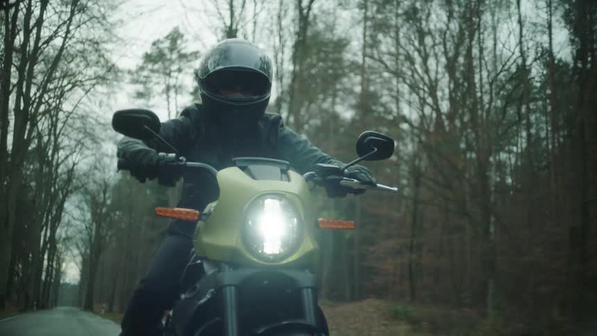 A cyclist wearing a black leather jacket and a helmet is riding a motorcycle on a road through the woods