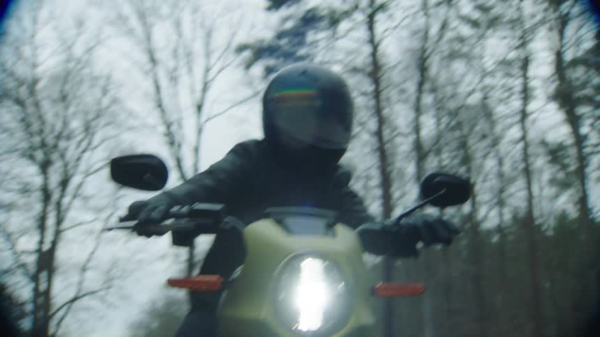 A cyclist wearing a black leather jacket and a helmet is riding a motorcycle on a wet road through the woods