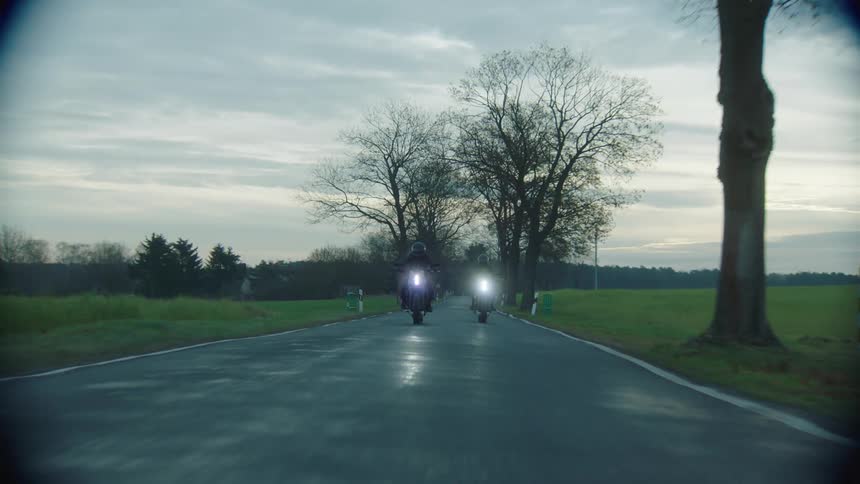 Two cyclists are riding motorcycles on a wet rural road