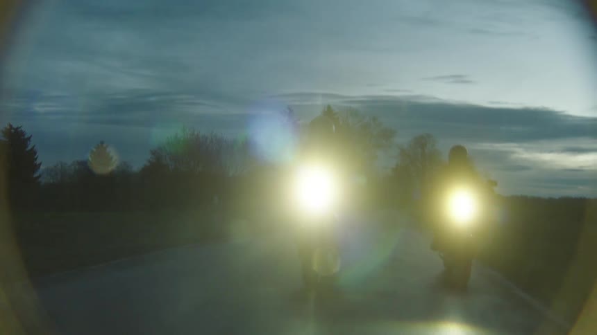 A pair of motorcyclists travel along a road through the countryside in the evening