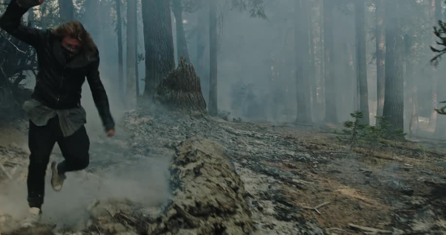 Young man wearing a balaclava runs over the ashes and burnt trees on a forest