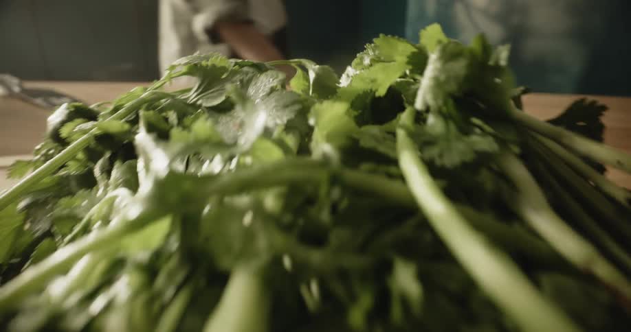 A person walks in the background where a bunch of cilantro sit on a cutting board