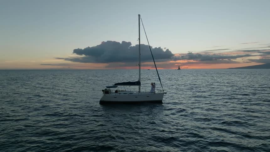 A couple standing over a sailboat in the ocean hugs at evening