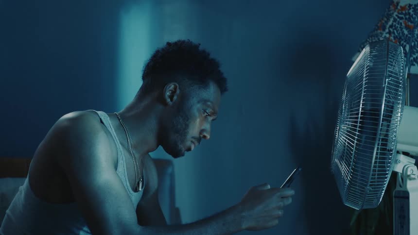 Young man sitting in front of a fan in the bedroom uses a smartphone