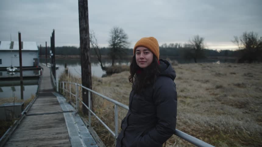 Woman smiles while leaning against rail on dock leading past buildings and field