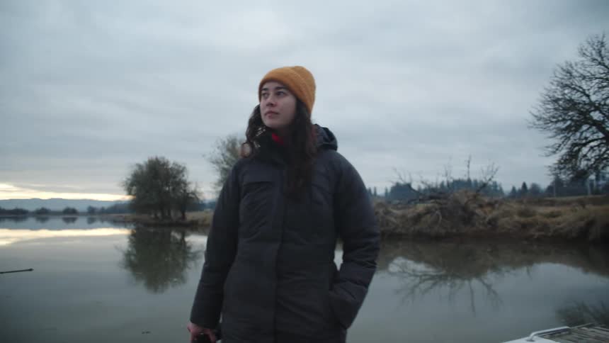 A woman is standing outside on a dock by a lake and holding a camera