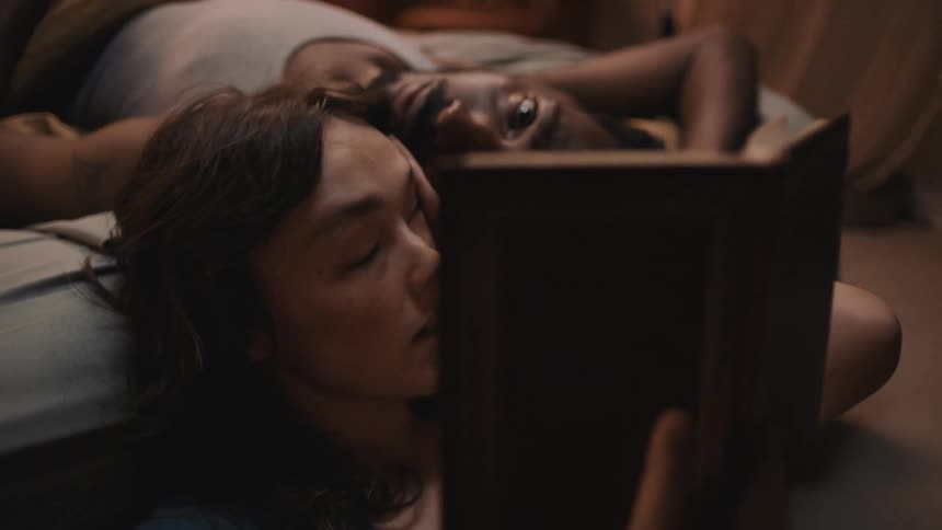 A man lays in bed as a woman sits nearby and reads