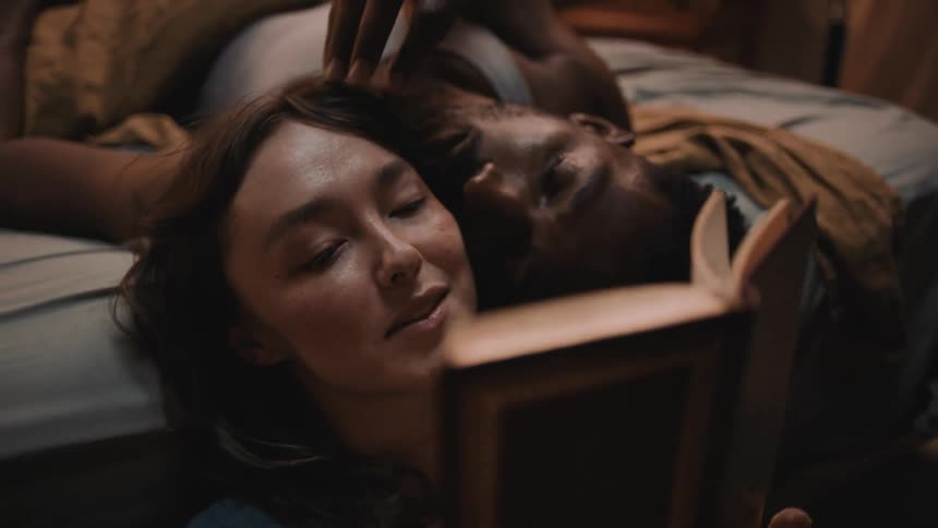 Woman reclined on the bed reads a book to her couple in the bedroom