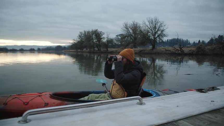 A woman is sitting in a canoe in a lake near the dock looking around with binoculars