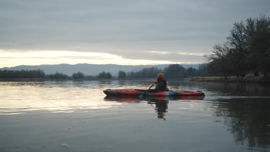 A woman is rowing a conoe across a lake to the forest on the other side and there are mountains in the distance