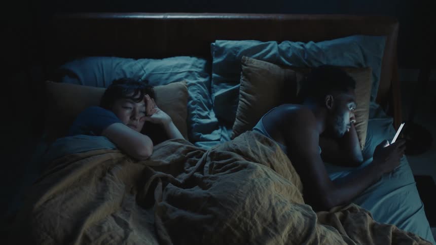 Woman sleeping in bed hits man with a pillow when she is woken by his smartphone screen light