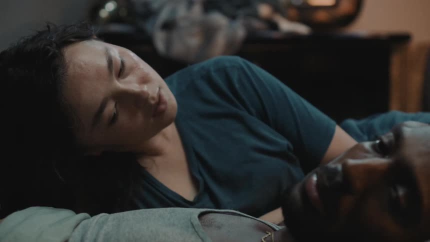 A man is lying in bed looking away from his wife as she looks at him with concern