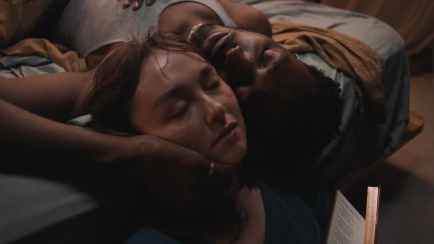 A woman holding a book leans against a bed and sleeps as a man in the bed caresses her face
