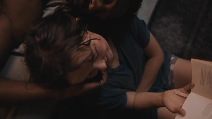 A man lies in bed and rests his head against a women sitting by the bed and caresses her face as she reads a book